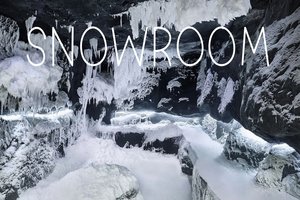 Wnętrze lodowej jaskini lub pomieszczenia typu snowroom z czarnymi skałami pokrytymi grubą warstwą śniegu i wiszącymi soplami lodu. Na górze obrazu widnieje duży, biały napis „SNOWROOM”.